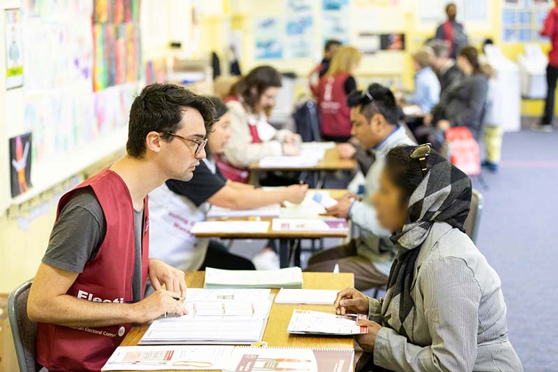 A VEC worker sits at a table across from a voter and looks up the voter's name on the roll. 