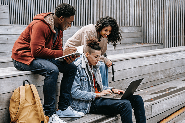 Three students sitting on steps, one is typing on a laptop and the others are looking over his shoulder. 