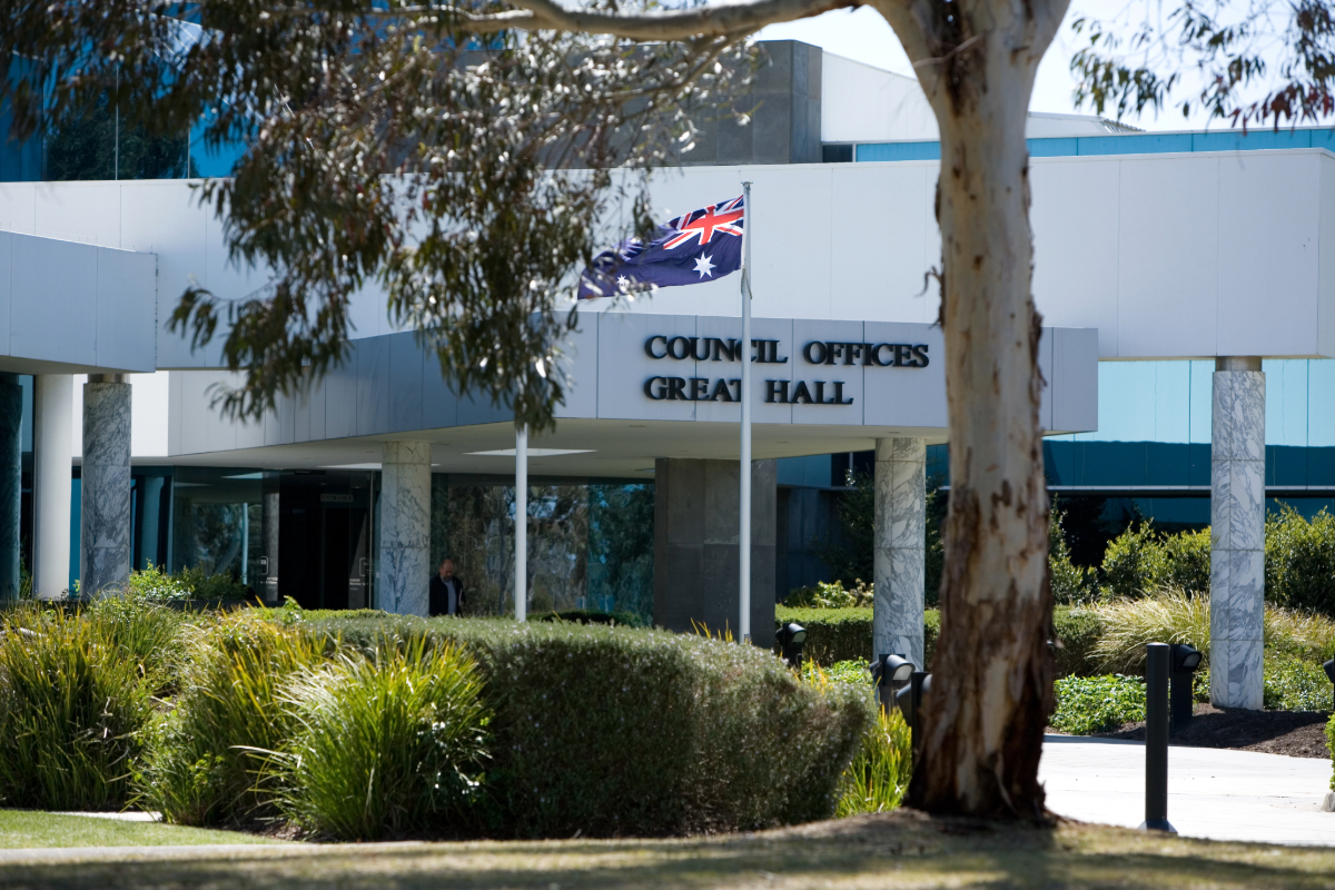 A white building with marble pillars and a sign that reads 'Council Offices Great Hall'. There is a large gumtree, hedges, grass and an Australian flag.