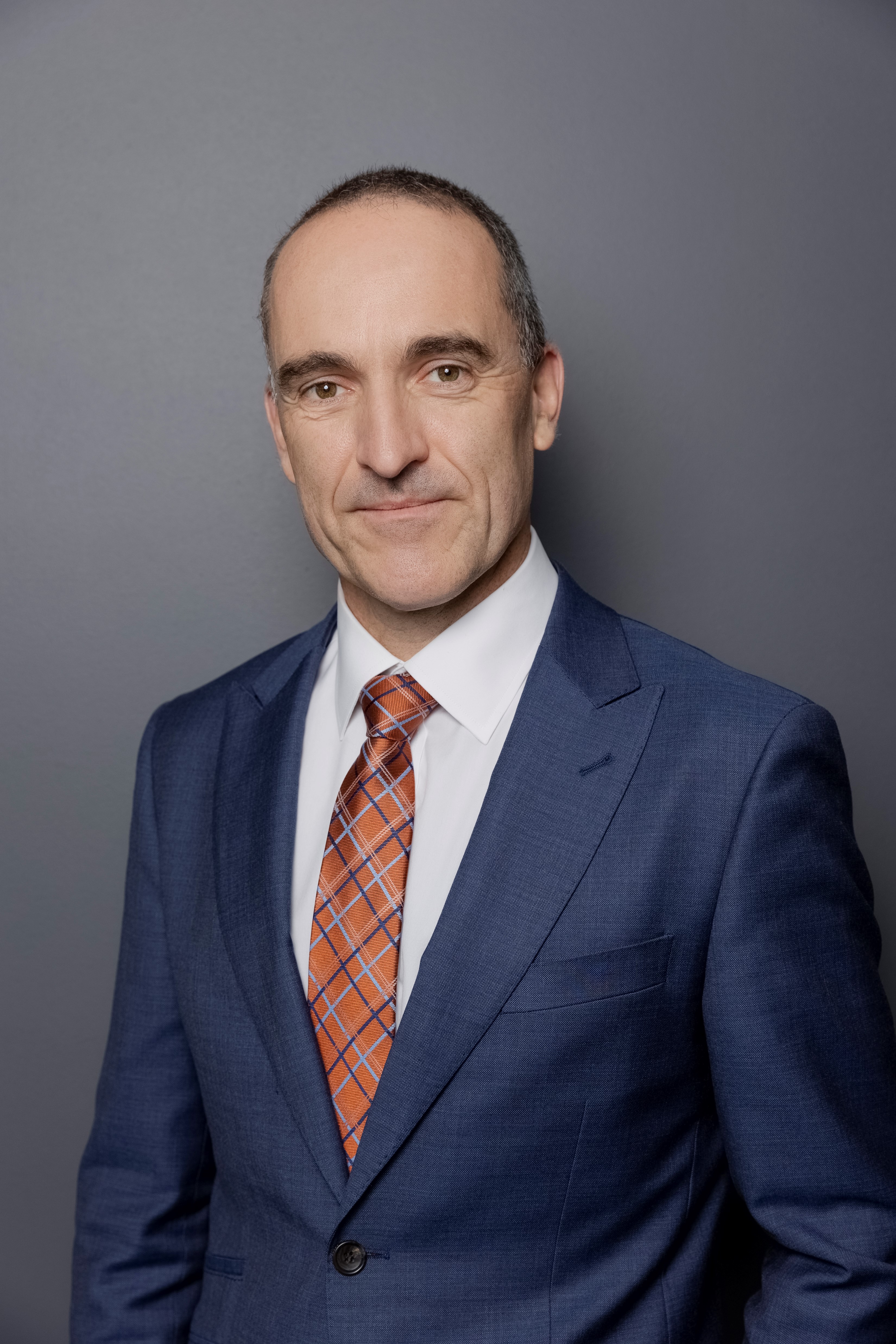Sven Bluemmel A professional portrait of the Victorian Electoral Commissioner, Sven Bluemmel, in a navy blue suit, white shirt, and orange plaid tie, standing against a plain grey background.