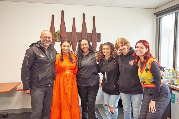 A group of people smiling for the camera. They are standing in front of a display of traditional Aboriginal weapons.