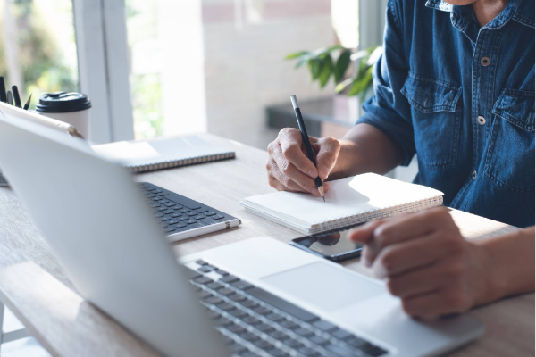 Person sitting at a desk using a laptop and writing notes in a notebook, with a phone and coffee cup nearby.