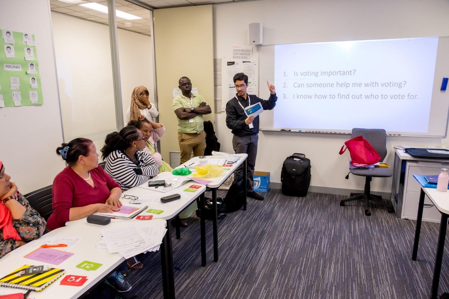 image of a classroom with a presentation on a whiteboard at the front of the room. students are facing the whiteboard. a VEC democracy ambassador is teaching at the whiteboard.