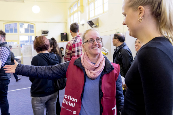 A smiling woman working at a voting centre is showing another woman to a voting booth. She is wearing a maroon vest that says 'Election official'.