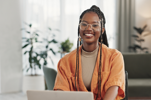 Person sitting at a desk and smiling. They are in front of a laptop screen in a bright indoor space with plants in the background.